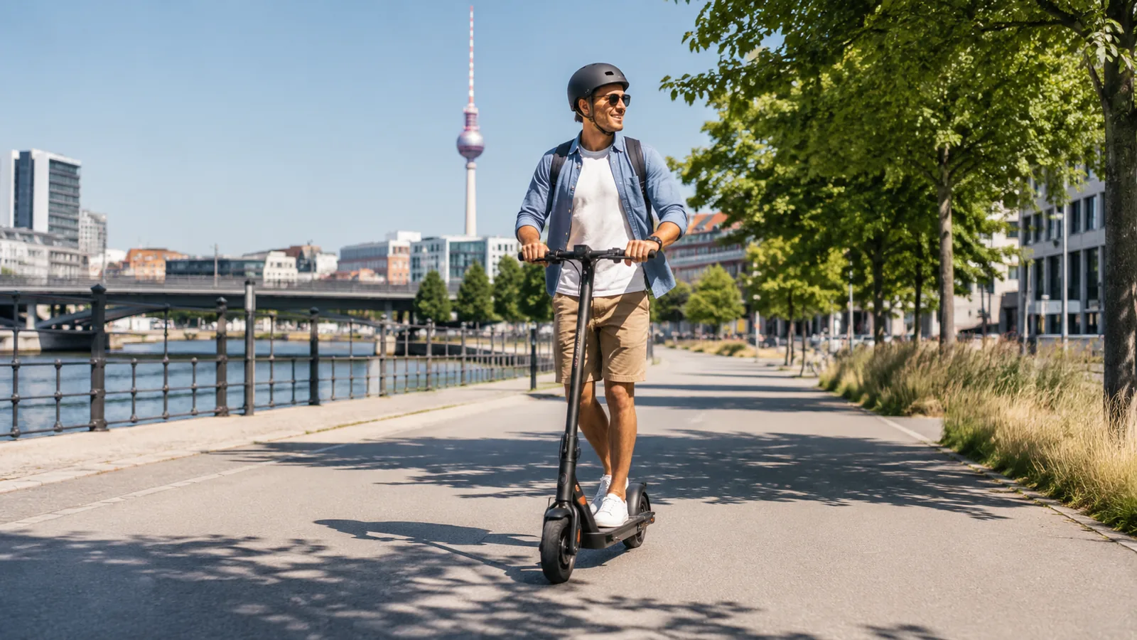 Mann mit Helm fährt an einem sonnigen Sommertag mit dem E-Scooter an der Uferpromenade in Berlin