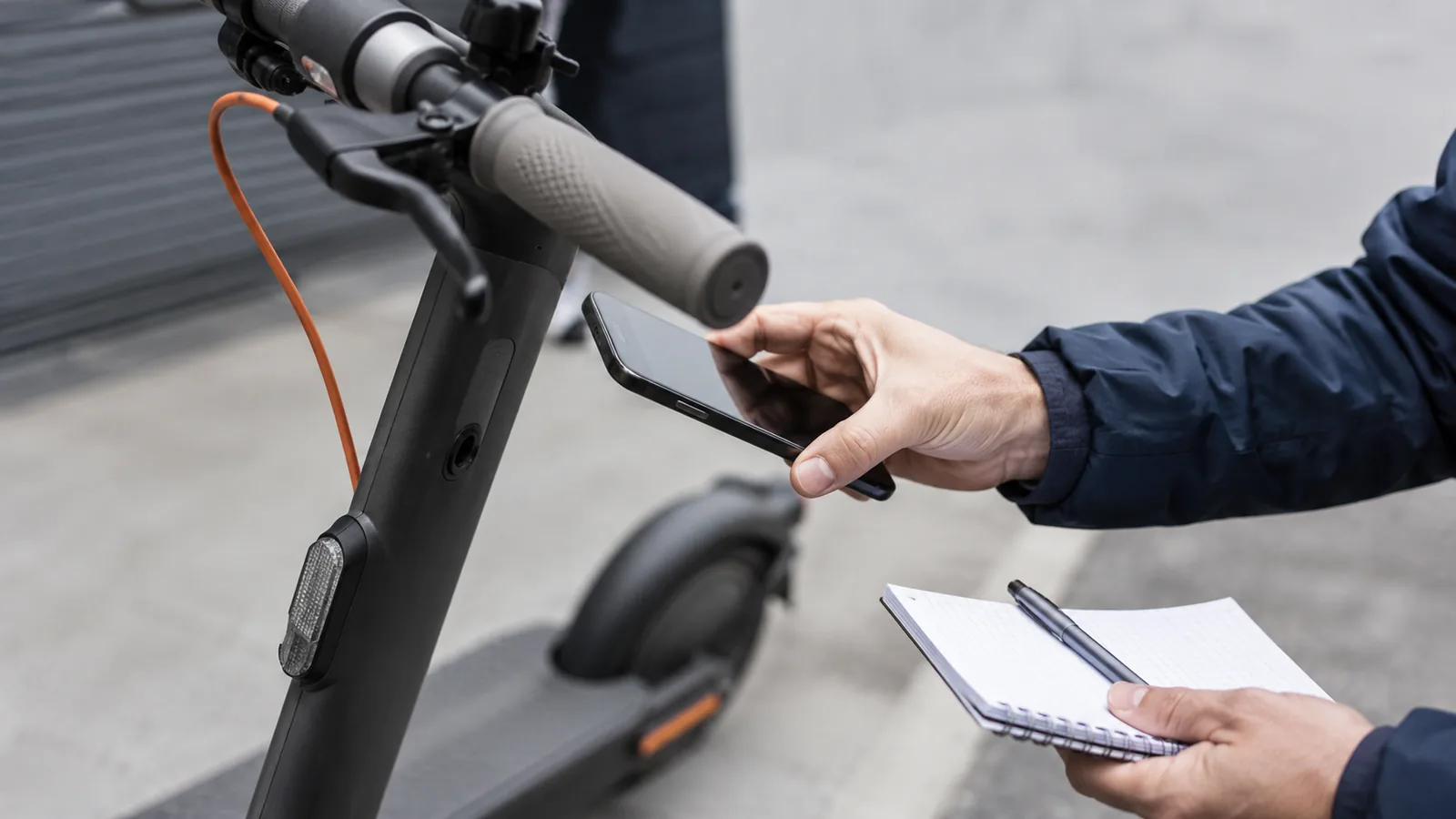 Person prüft am E-Scooter-Lenker mit Smartphone und Notizblock die Seriennummer, keine lesbaren Zeichen